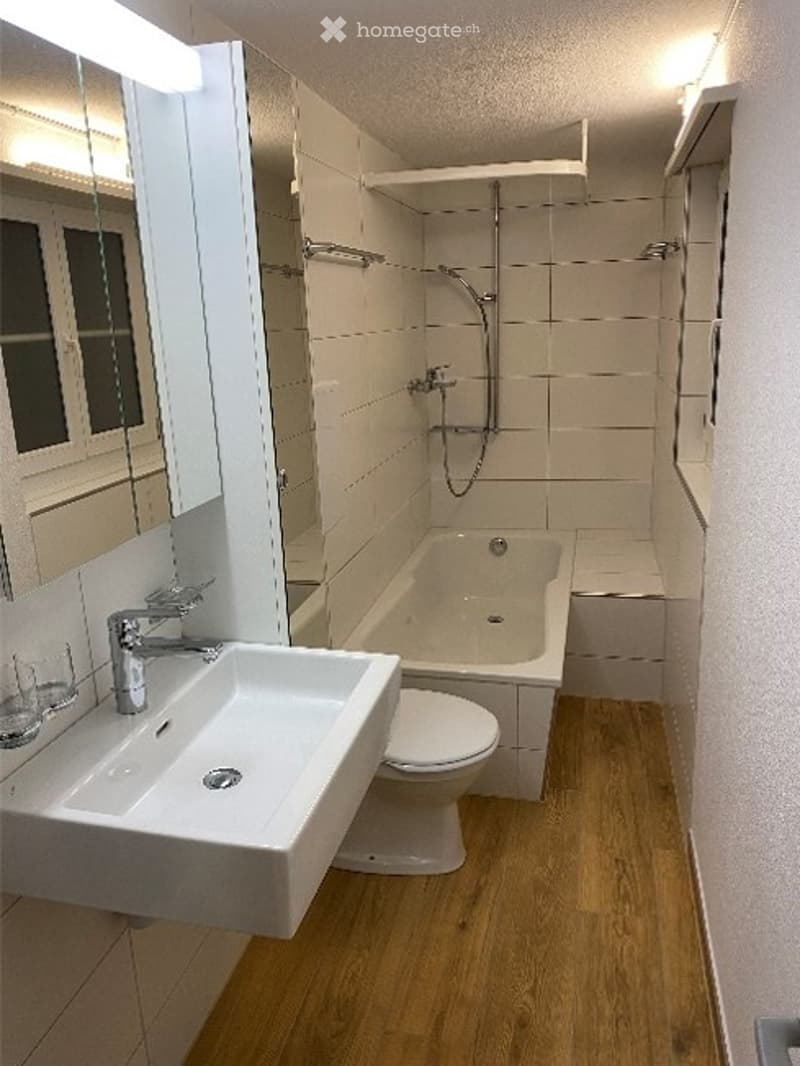 Modern bathroom with white tiles, wooden flooring, a sink with a chrome faucet, a bathtub, a toilet, a shower, and a mirrored cabinet.