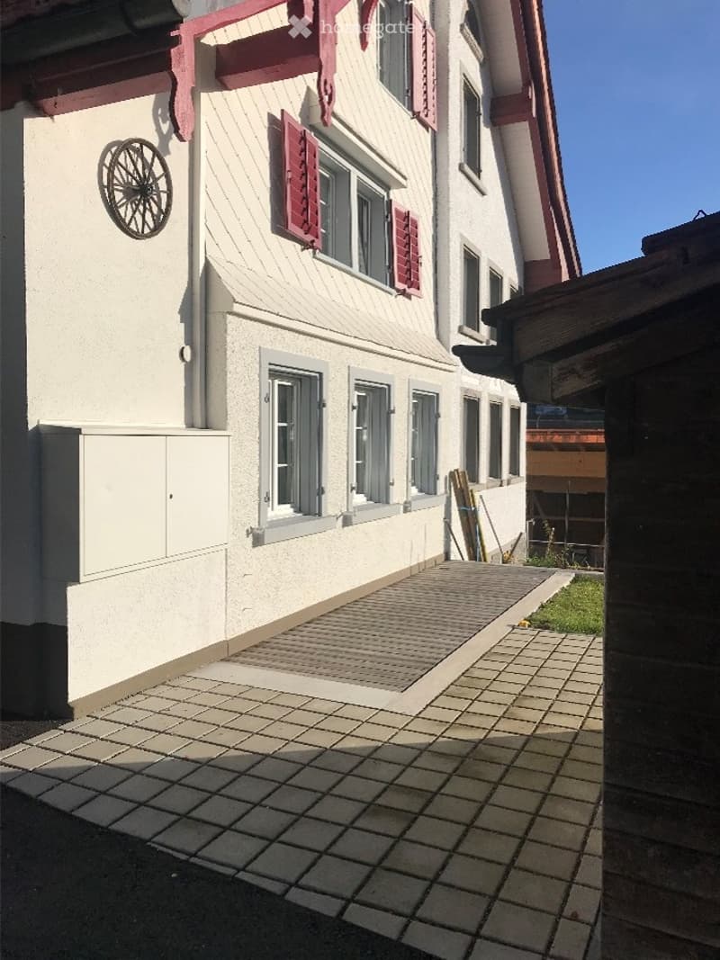 Two-story building with white walls, red shutters, and a tiled entrance area. Decorative wheel design on the wall.