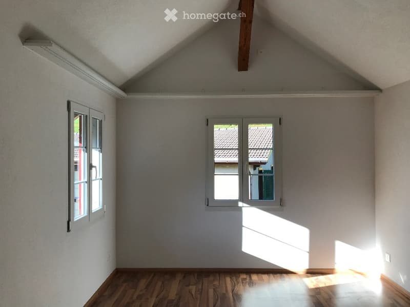 Empty attic room with wooden flooring, white walls, and two windows allowing natural light to enter.