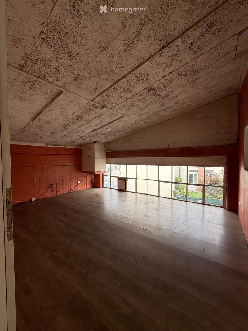 Empty room with exposed wooden ceiling, concrete floor, large windows, and red wooden paneling on one wall.