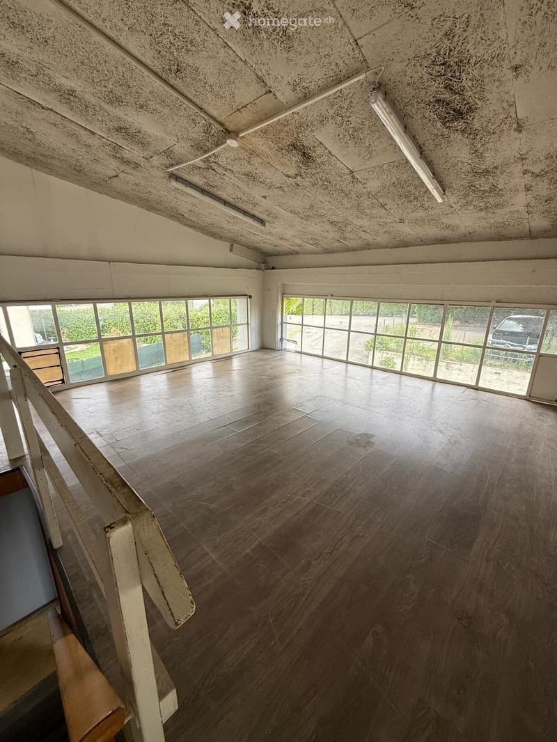 An empty room with wooden flooring, large windows, and a staircase leading downwards. The ceiling has exposed beams and lighting fixtures.