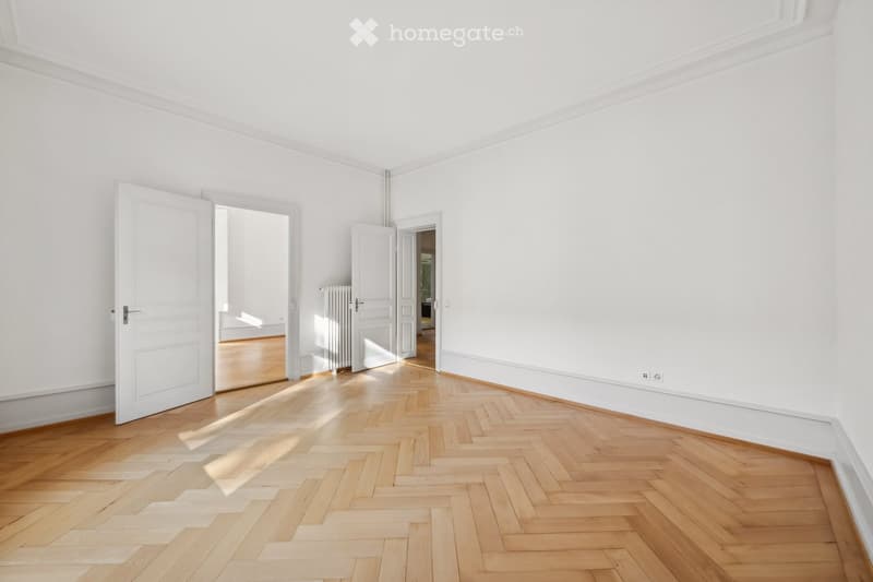 Empty room with white walls, wooden herringbone floor, and open doors leading to other rooms.