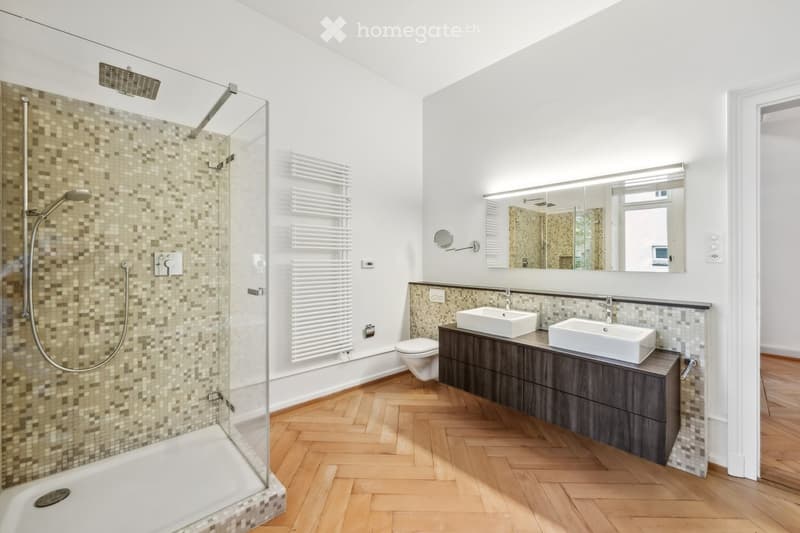 Modern bathroom with a glass-enclosed shower, double sink vanity, toilet, and a large mirror above the sinks. The floor is wooden, and the walls feature a mix of tiles and white paint.