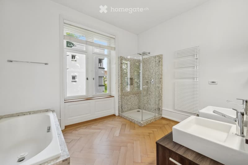 Modern bathroom with a white bathtub, double sink vanity, glass-enclosed shower, and a radiator. The room features wooden flooring and large windows with blinds.