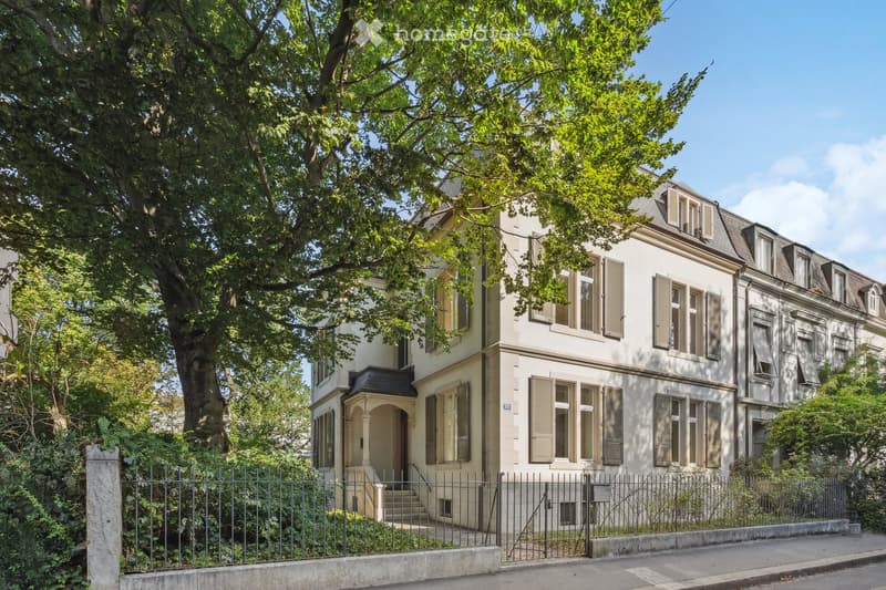 A three-story residential house with light-colored walls, multiple windows with shutters, and a small entrance porch. The house is surrounded by greenery and a metal fence.