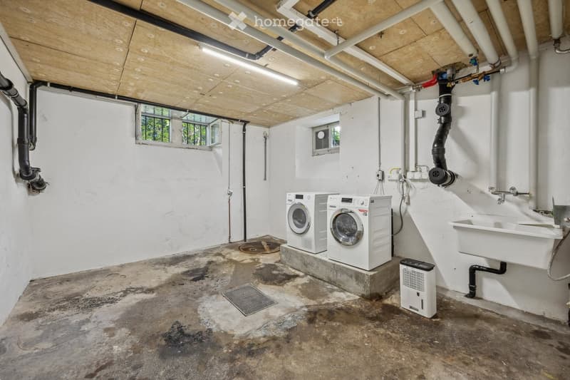 Utility room with white washing machine, dryer, sink, and air conditioner. Exposed pipes and concrete floor.