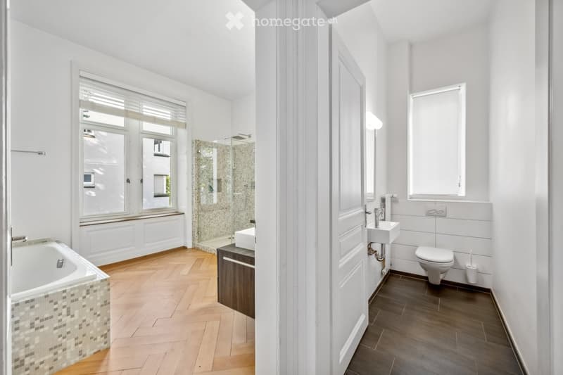 Modern bathroom with a bathtub, sink, toilet, and shower area. Features large windows, wooden flooring, and white walls.