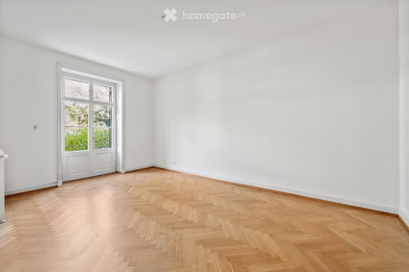 Empty room with white walls, wooden herringbone flooring, and a large window with a door leading outside.