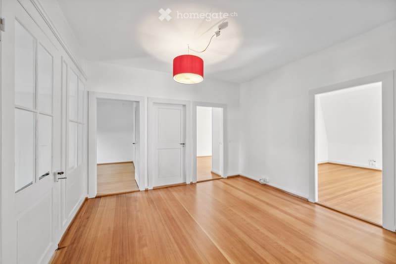 Empty hallway with white walls, wooden flooring, and several doors leading to other rooms. A red pendant light hangs from the ceiling.