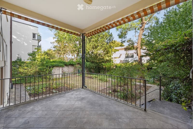 Balcony with tiled floor, metal railing, and covered awning, overlooking a lush green garden area with trees and shrubs.
