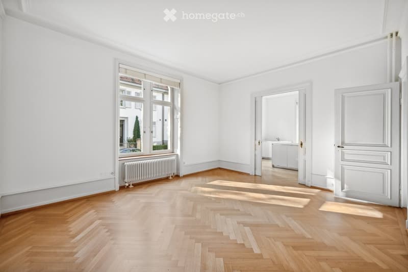 Empty room with white walls, wooden herringbone flooring, large window with radiator below, and two open doors leading to adjacent spaces.