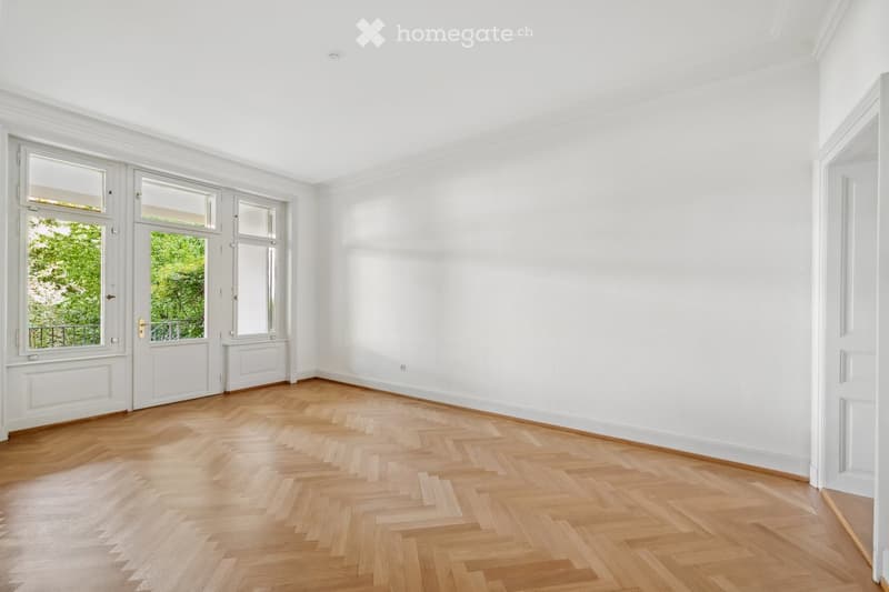 Empty room with white walls, wooden herringbone flooring, large windows, and a door leading to a balcony.