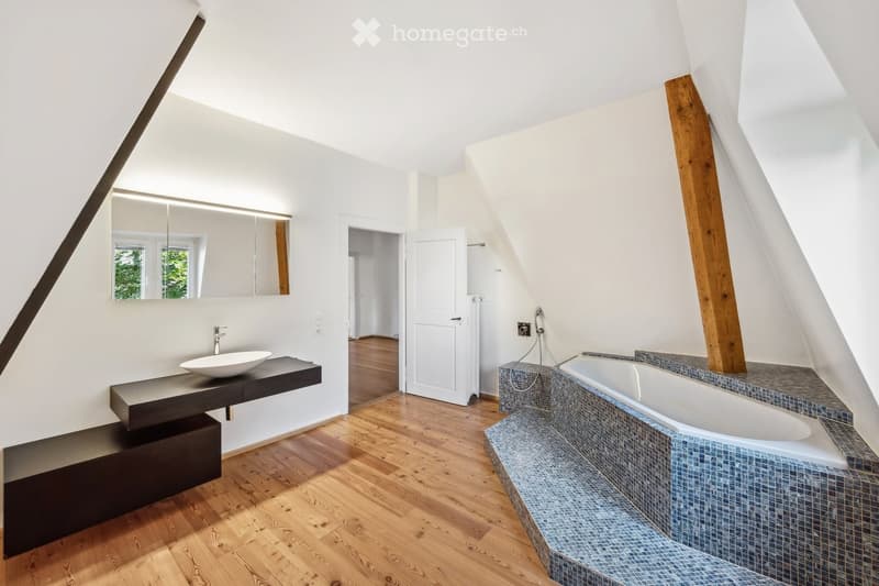 Modern bathroom with a sleek black sink, a bathtub with blue tiles, and a large mirror above the sink. The room features wooden flooring and white walls with exposed wooden beams.