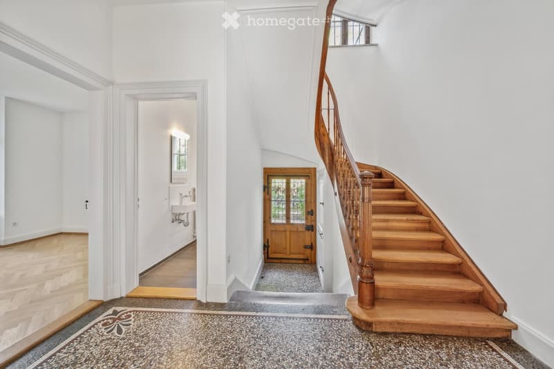 Elegant hallway with wooden staircase, polished wooden floors, white walls, and a small sink area visible in the background.