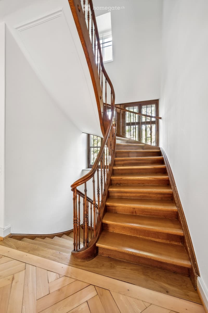 Spiral staircase with wooden steps and handrail, white walls, and large windows allowing natural light to enter.
