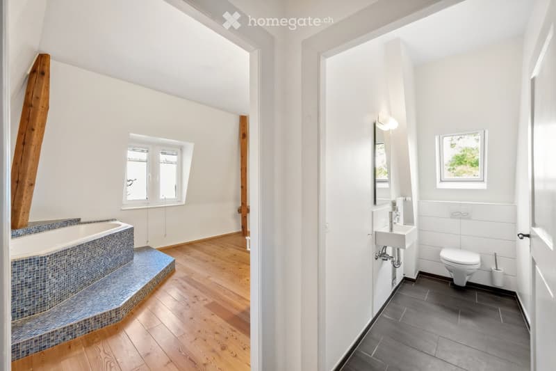 Modern bathroom with a bathtub, sink, and toilet. The space features wooden flooring, white walls, and large windows providing natural light.