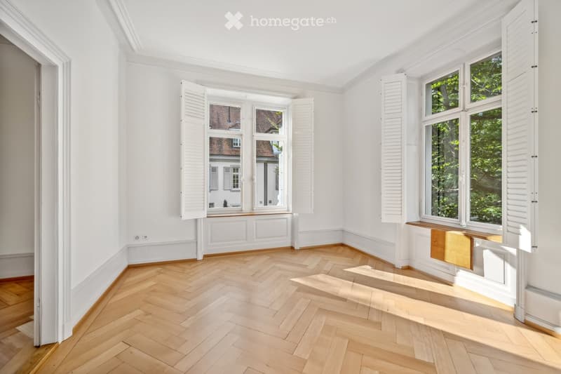 Empty room with white walls, herringbone wooden flooring, large windows with shutters, and a small wooden bench under one window.