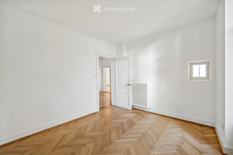 Empty room with white walls, wooden herringbone flooring, a small window, and an open door leading to another room.