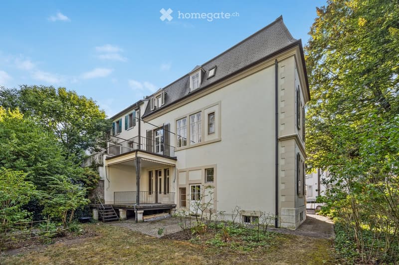 A two-story house with a raised ground floor, featuring a balcony, multiple windows, and a staircase leading to the entrance.