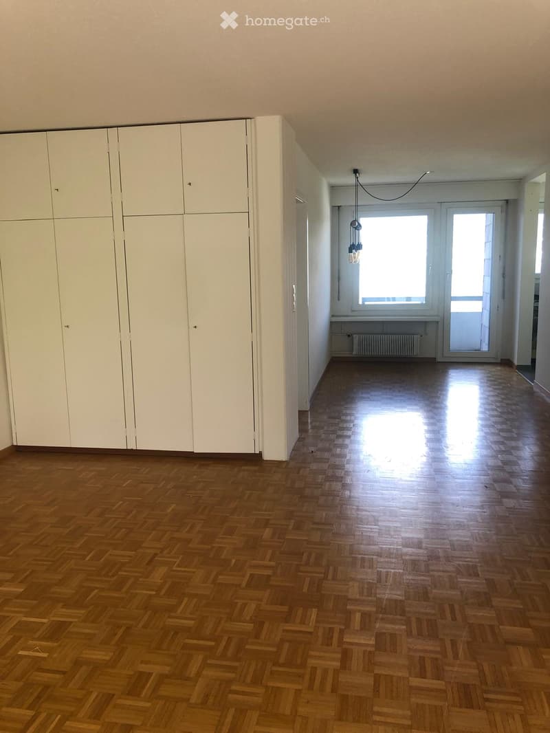 Empty room with wooden herringbone flooring, white built-in wardrobes, and a large window with a view of the outside.