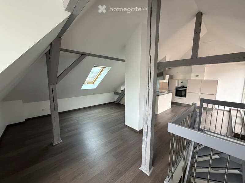 Bright, open hallway with wooden flooring, white walls, and a modern staircase leading upwards. Features include exposed wooden beams, a skylight window, and a small built-in shelf on the left side.