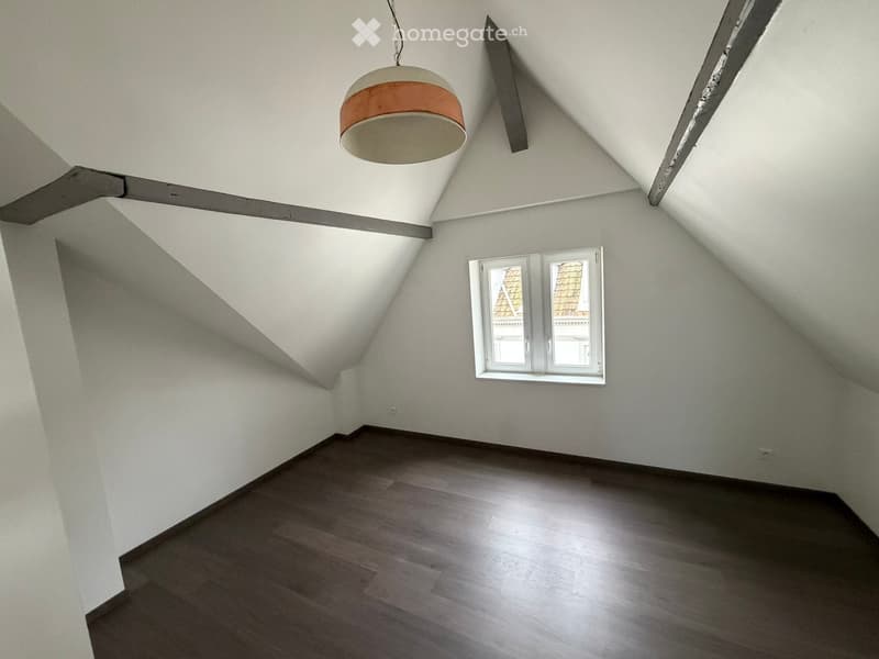 Empty attic room with wooden flooring, white walls, and a single window offering a view of the roof. A modern pendant light hangs from the slanted ceiling.