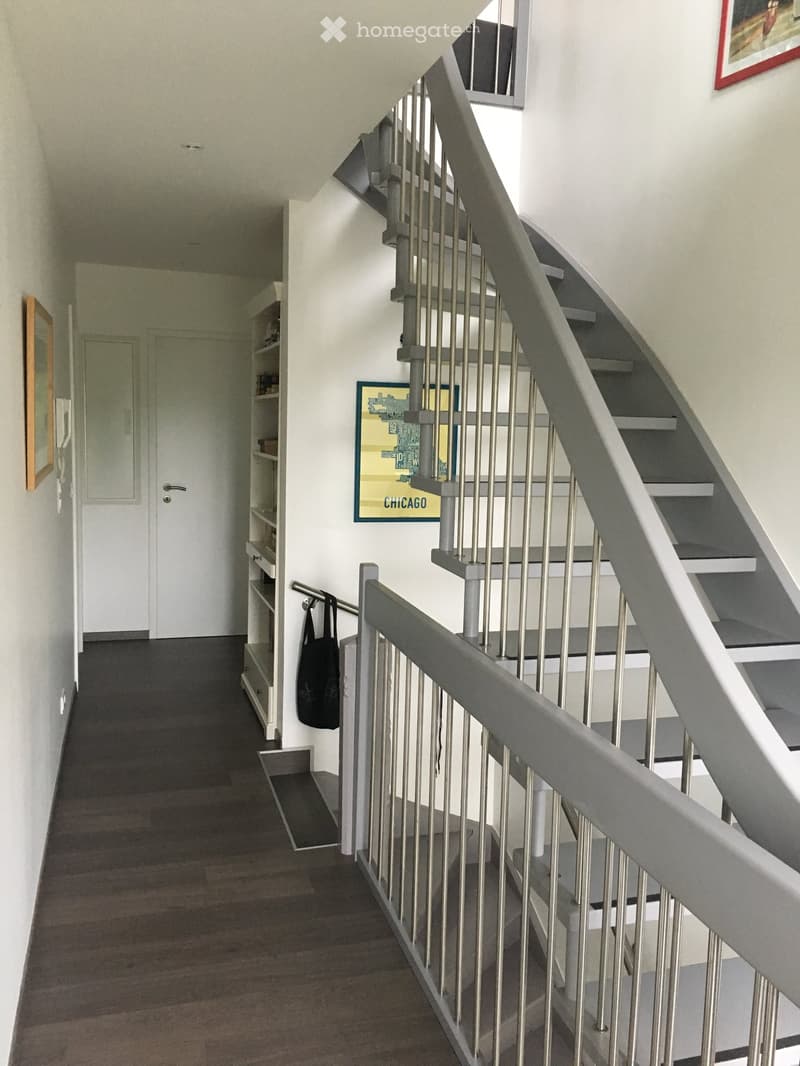 Modern hallway with wooden flooring, white walls, a staircase with metal railings, a shelf with books, and framed artwork on the walls.