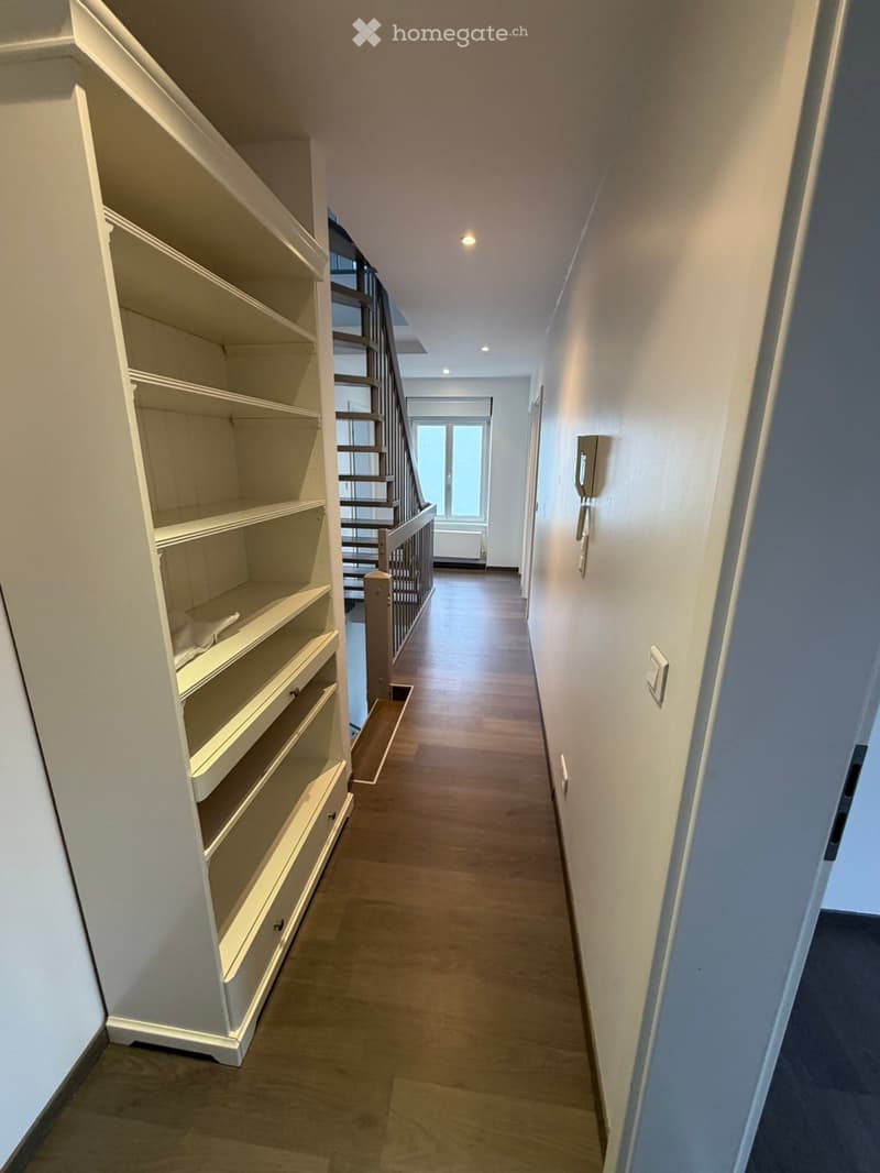 Empty hallway with wooden flooring, white walls, built-in shelving unit on one side, and a staircase leading upwards.