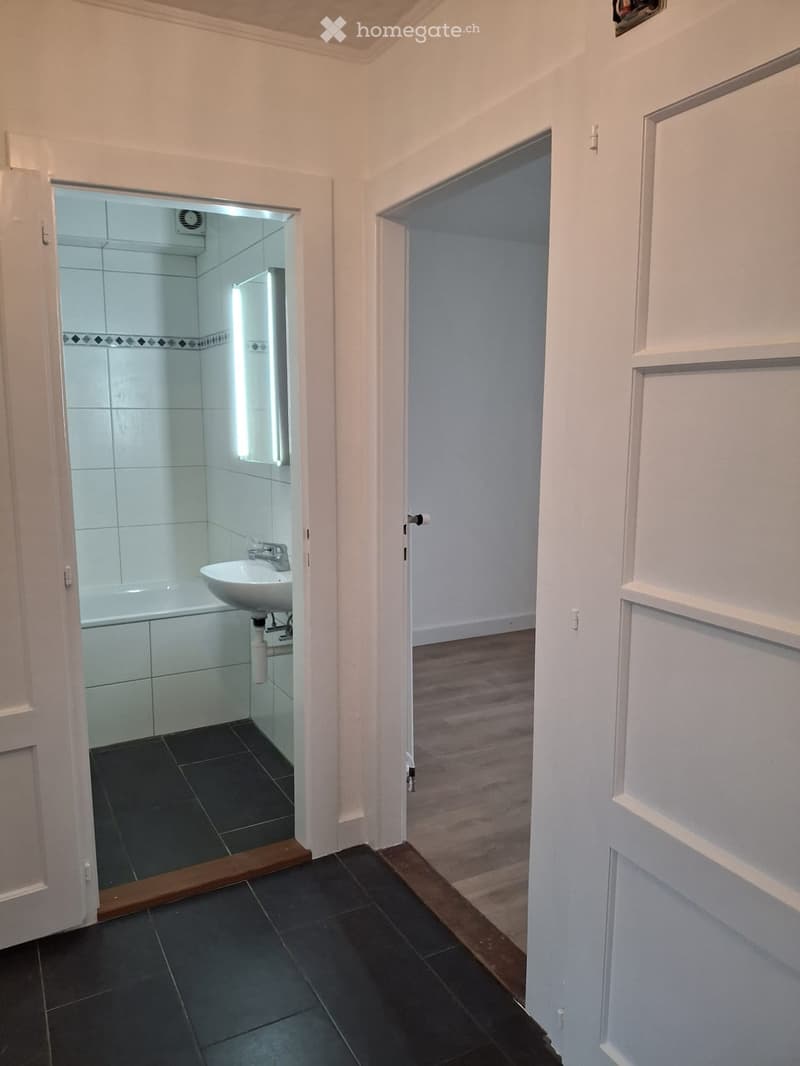 Bathroom with a sink and bathtub visible through an open door, featuring dark tiled floors and white tiled walls.