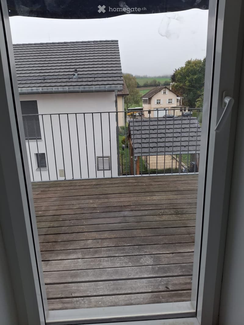 Wooden balcony with metal railing, overlooking a residential area with houses and greenery.
