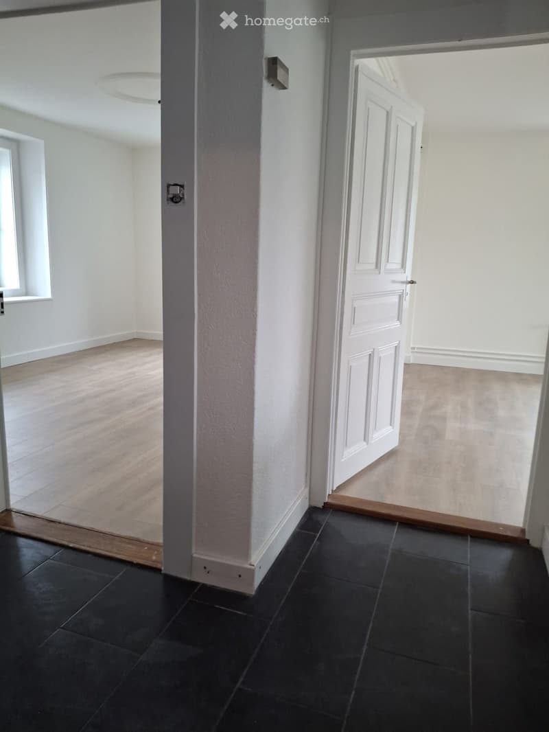 Empty hallway with white walls, black tiled floor, and wooden flooring in adjacent rooms. Features a central column and open doors leading to other spaces.