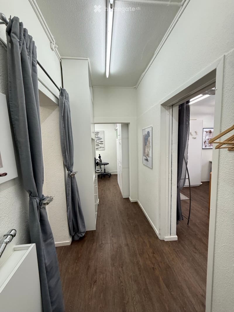 A narrow hallway with wooden flooring, white walls, and grey curtains on both sides. The hallway leads to an open doorway revealing a room with a chair and artwork on the walls.