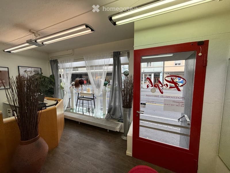 A modern, well-lit room with a wooden floor, large windows with sheer curtains, a red door, a stool, a laptop on a counter, and decorative vases.