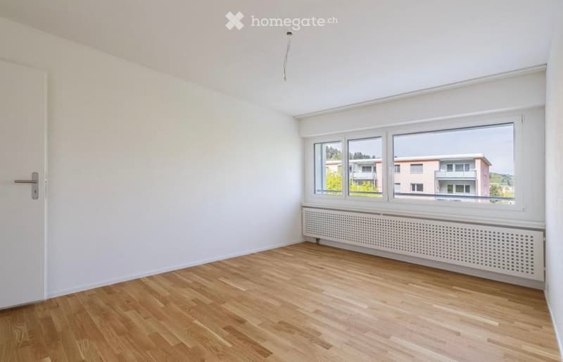 Empty room with wooden flooring, white walls, and large windows offering a view of neighboring buildings.