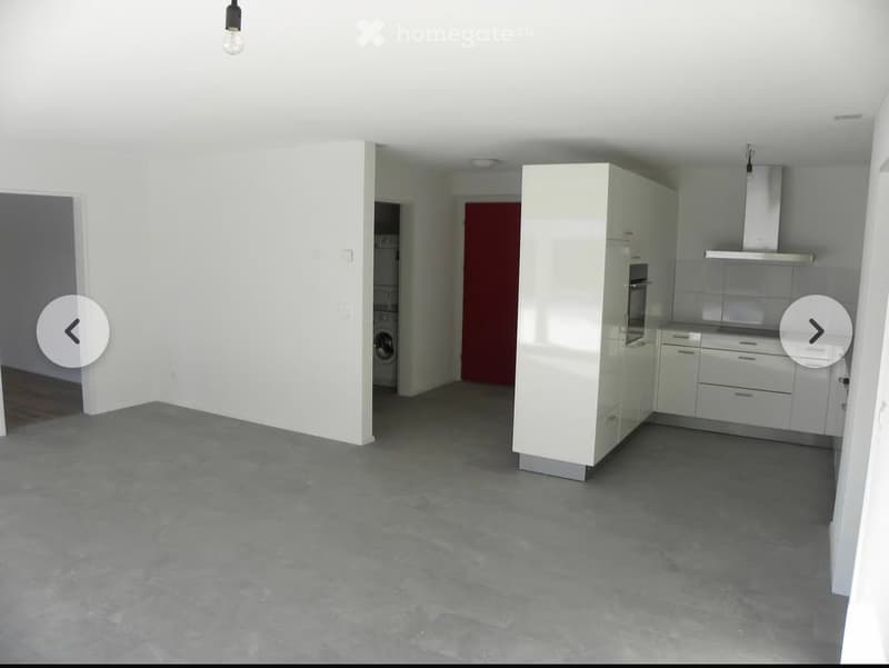Empty room with grey concrete floor, white walls, and a white kitchen area with cabinets, a washing machine, and a range hood in the background.