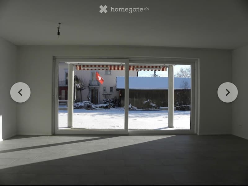 Empty room with large sliding glass doors leading to a snowy outdoor area, featuring a Swiss flag and a covered patio.