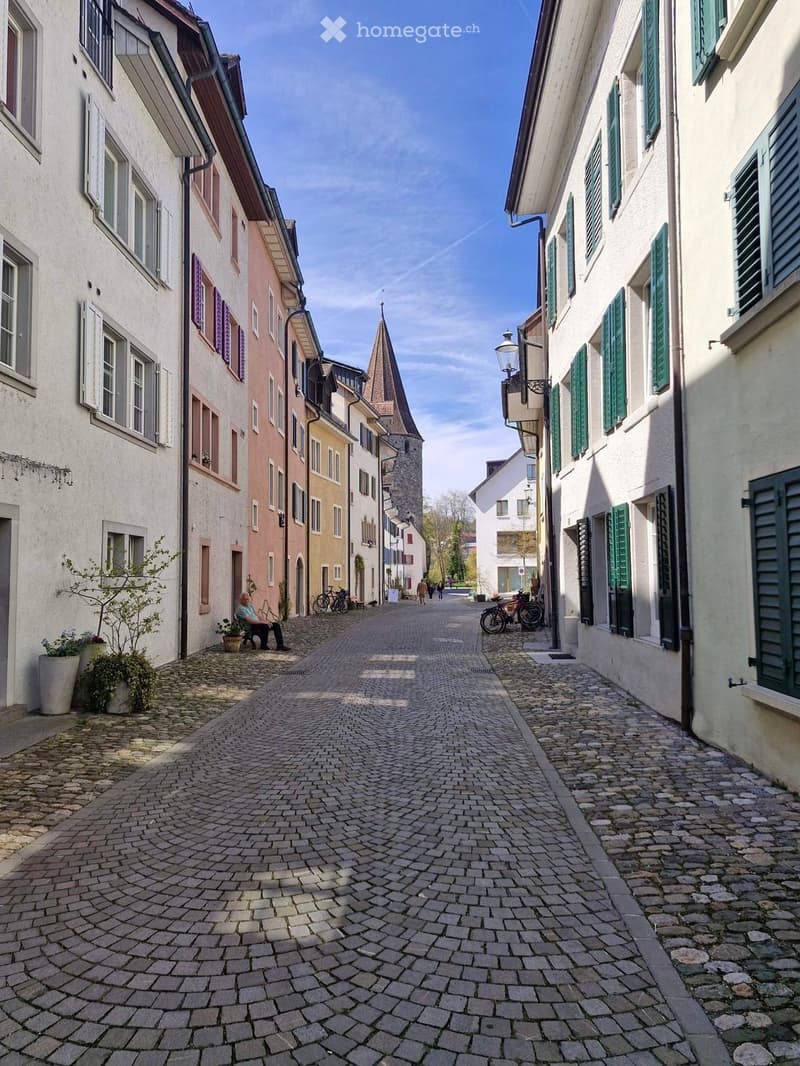 A narrow cobblestone street lined with colorful buildings, featuring a bench, potted plants, bicycles, and a distant church tower.