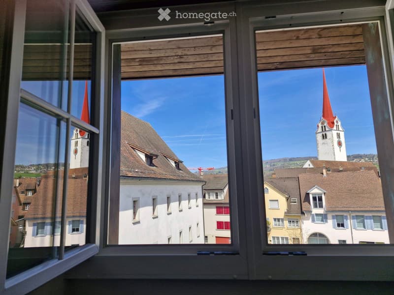View from a window showing a town with traditional buildings, a church tower with a red spire, and a clear blue sky.