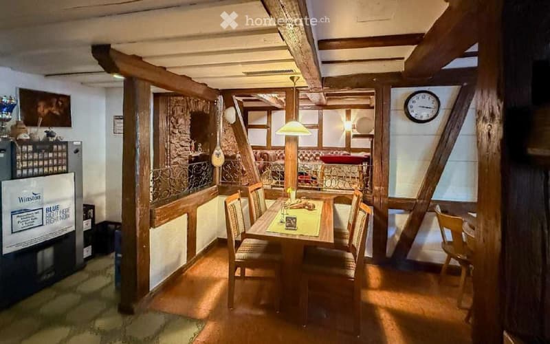 A cozy dining area with a wooden table and chairs, a clock on the wall, and a refrigerator with a sign on it. The room features exposed wooden beams and warm lighting.
