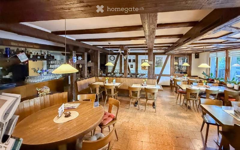 Cozy restaurant interior with wooden tables and chairs, pendant lights, and a bar area with shelves and glassware.