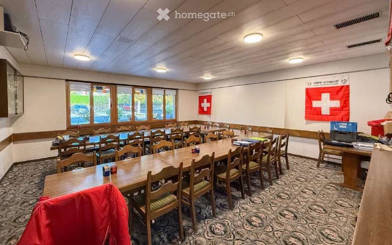 A large conference or meeting room with multiple tables and chairs arranged in rows, large windows, and Swiss flags on the walls.
