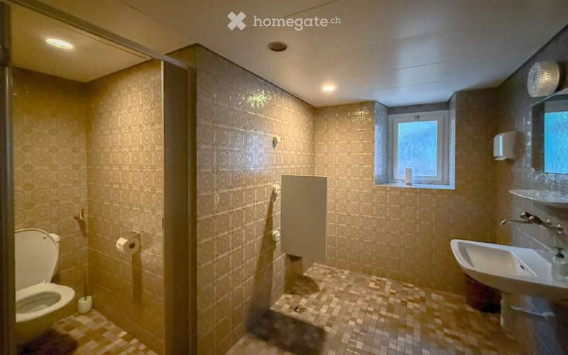 Modern bathroom with beige tiles, a toilet, a sink with a mirror, and a shower stall. The room features a small window and built-in storage shelves.