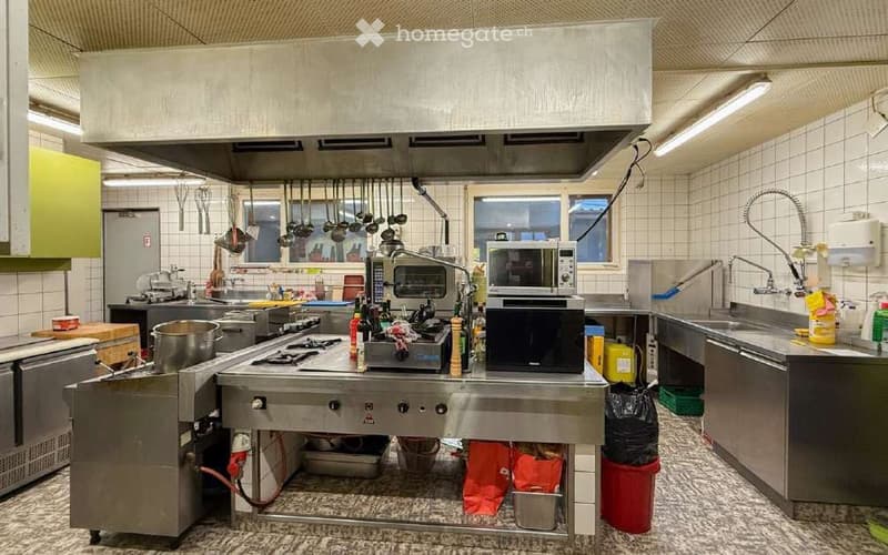 Commercial kitchen with stainless steel appliances, stove, microwave, sink, hanging pots and pans, and various kitchen tools.