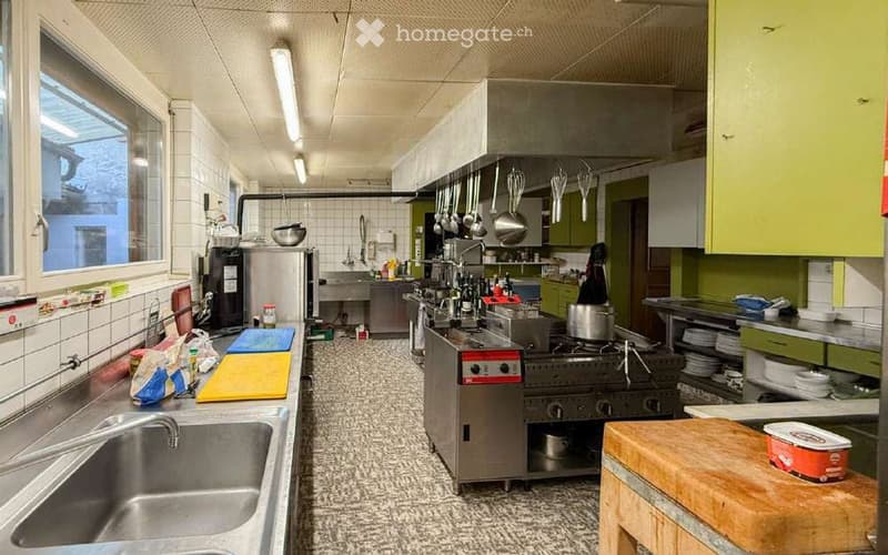 Commercial kitchen with stainless steel appliances, including a sink, stove, oven, and refrigerator. Pots and pans hang from the ceiling, and various kitchen tools and ingredients are visible on the counters.
