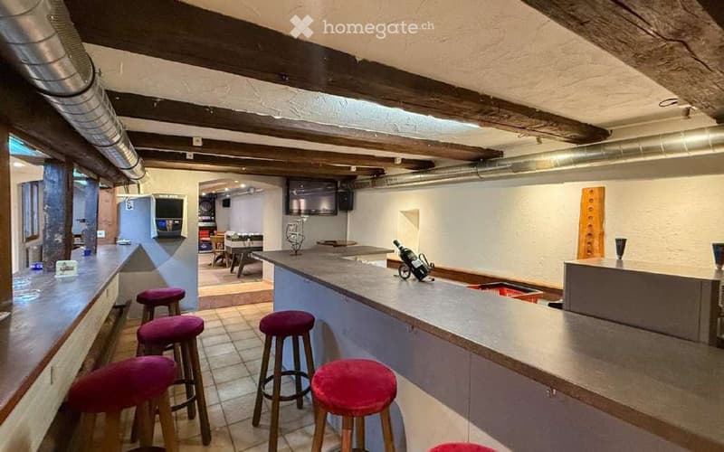 A cozy bar area with a wooden bar counter, red cushioned stools, a bottle on the counter, a TV screen mounted on the wall, and a small table with chairs in the background.