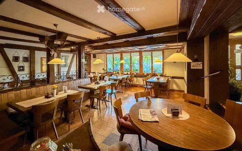 Restaurant interior with wooden tables and chairs, exposed wooden beams, hanging lamps, and large windows with a view of the garden.