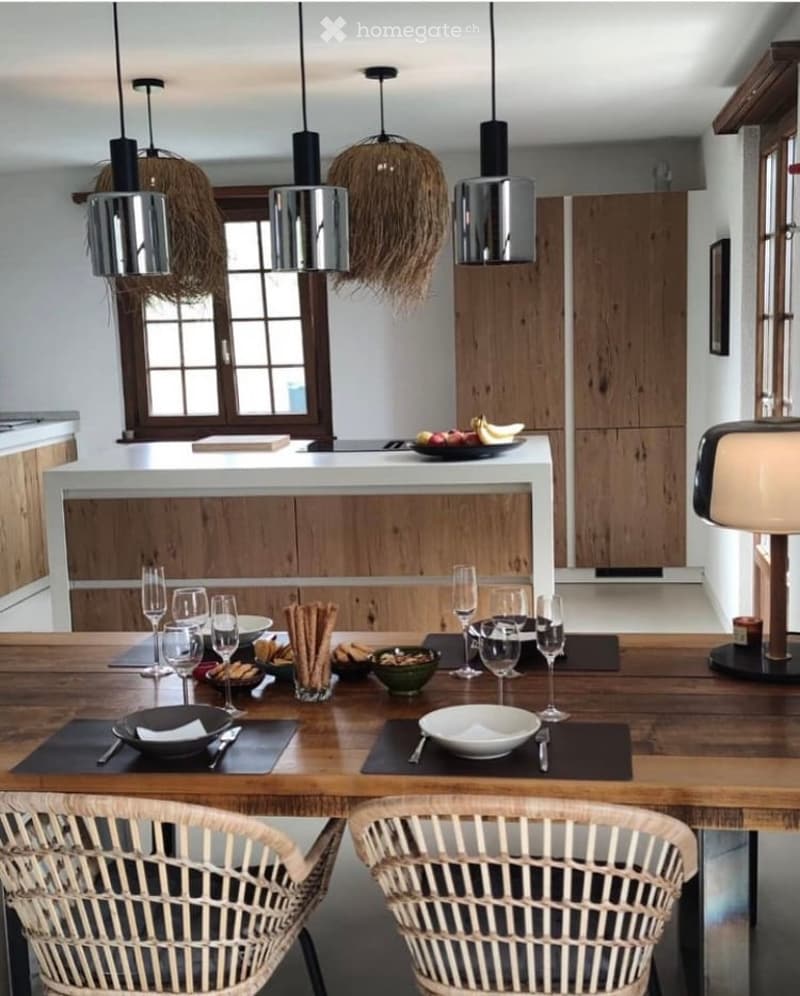 Modern kitchen with wooden cabinets, white countertops, and a dining area featuring a wooden table set with wine glasses, plates, bowls, and various food items. The space includes hanging pendant lights and wicker chairs.