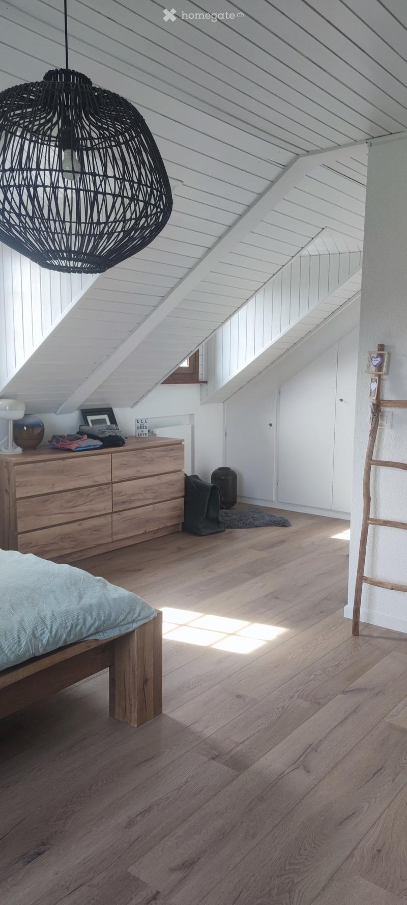 Bright attic bedroom with wooden flooring, slanted white ceiling, and large windows allowing natural light. Features a wooden bed, dresser with various items, a wicker pendant light, and a small rug.