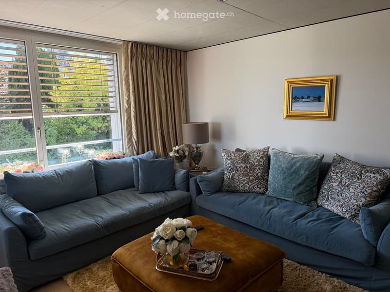 A modern living room with a blue sectional sofa, a wooden coffee table with a flower vase, a table lamp, and decorative cushions. Large windows with blinds offer a view of greenery outside.
