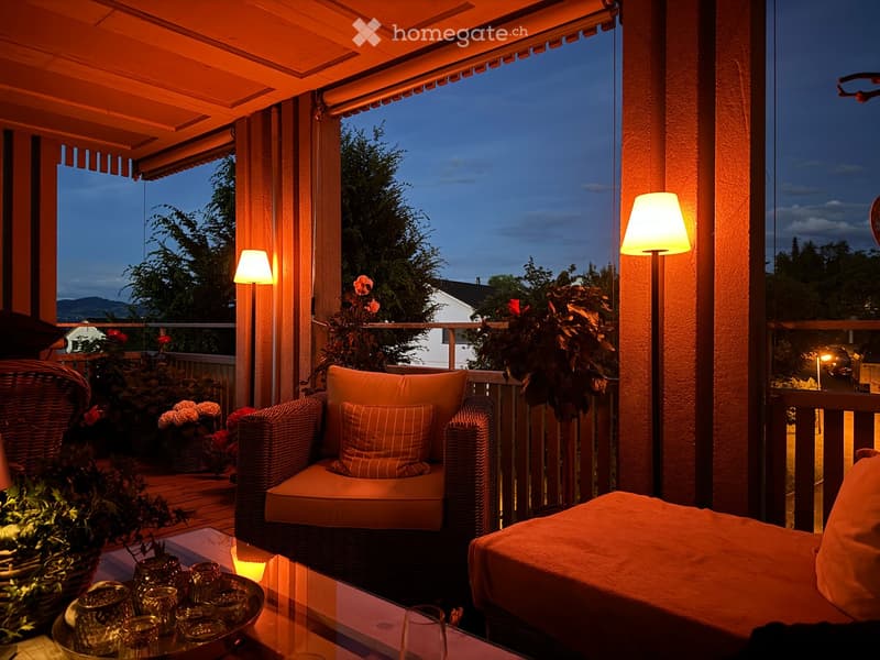 Well-lit balcony with wicker seating, cushions, glass table, and decorative plants, offering a scenic evening view.